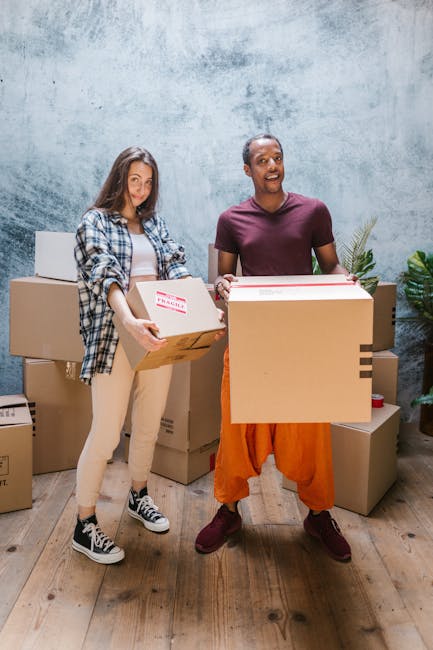 Two women are standing inside a spacious room during a home relocation process, surrounded by several large cardboard boxes sealed with red tape. One woman, on the left, holds a medium-sized box with