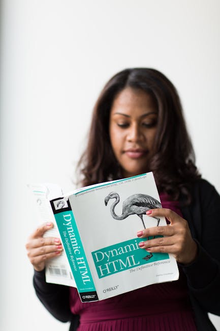 A woman with dark brown hair, dressed in a black blazer and maroon top, is reading a large book titled 'Dynamic HTML' by O'Reilly, which features a flamingo illustration on the cover. She is standing 